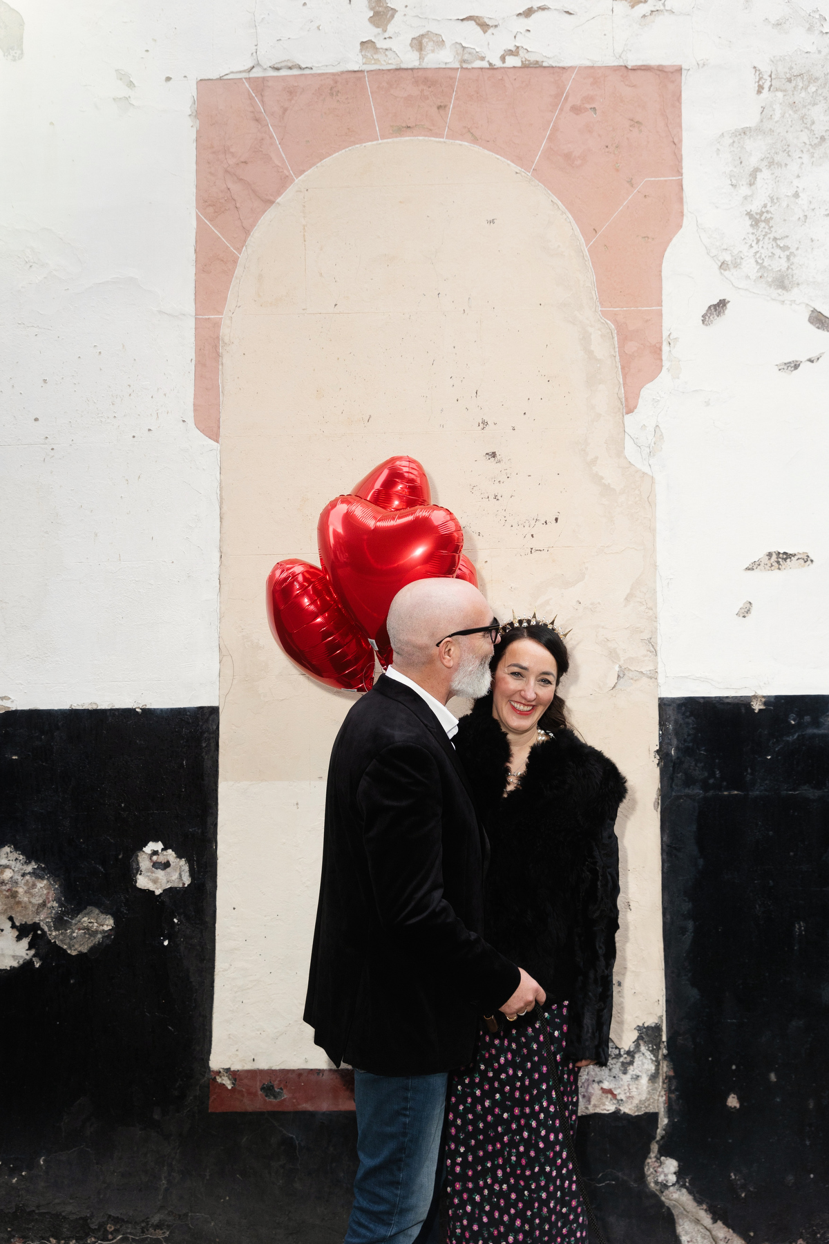 Bride and groom portrait with red heart balloons on rustic urban backdrop.